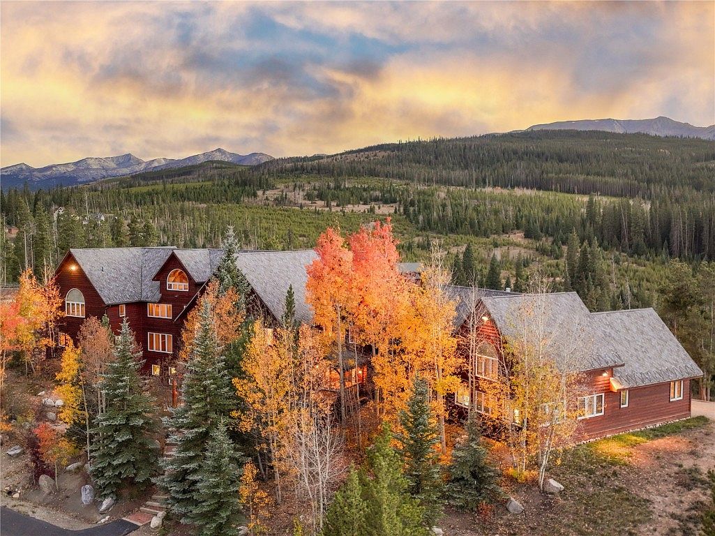 This is an eye-level, wide shot of a large, multi-sectioned log cabin home nestled among colorful autumn trees and evergreen pines. The home features multiple gabled roofs, numerous windows, and a warm, inviting exterior. In the background, forested hills and distant mountains are visible under a dramatic, colorful sky, creating a serene and picturesque setting.