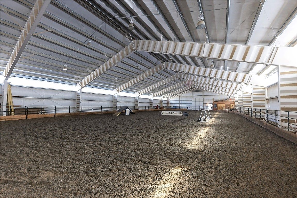 This is an interior shot of a large indoor riding arena. The arena features a sand or dirt floor, metal roofing and support beams, and fencing around the perimeter. There are various obstacles set up in the arena, suggesting it is used for training or practice.