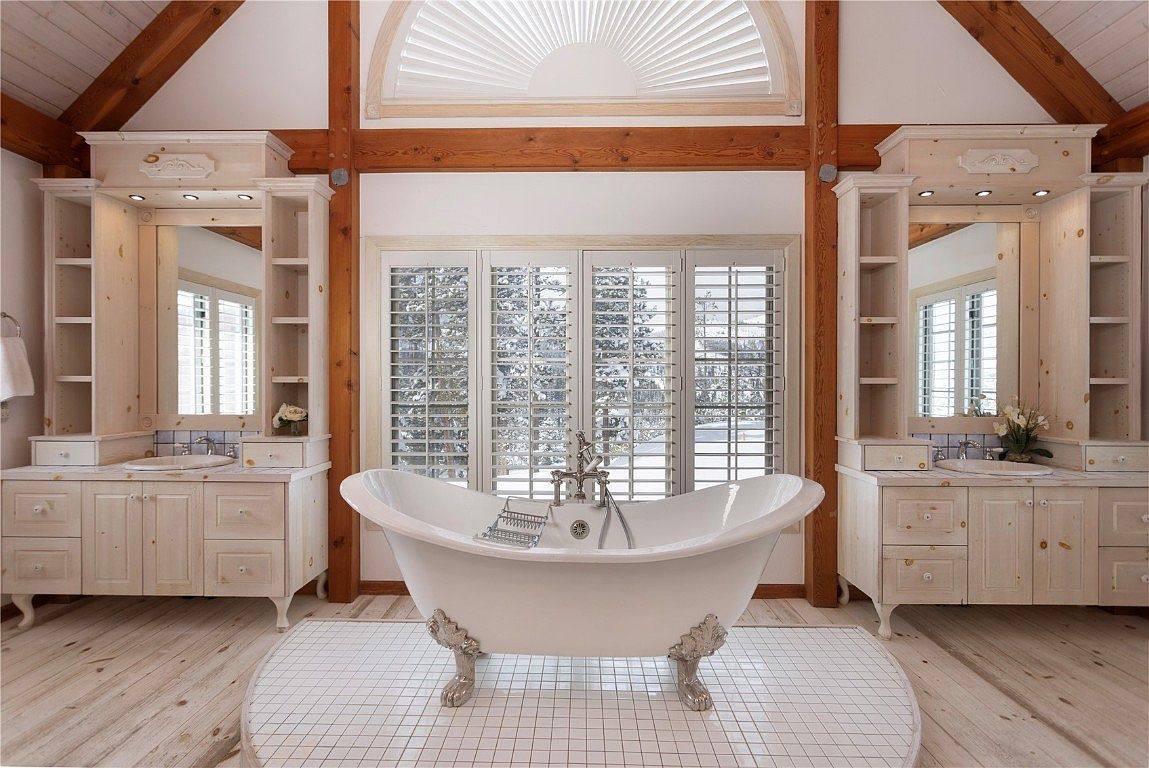 This is a bright and airy primary bathroom featuring a freestanding clawfoot bathtub as the focal point, positioned in front of a window with plantation shutters. The room is flanked by matching light wood vanities with built-in shelving and mirrors. The flooring consists of light wood planks and a circular tiled area around the tub, creating a luxurious and inviting spa-like atmosphere.