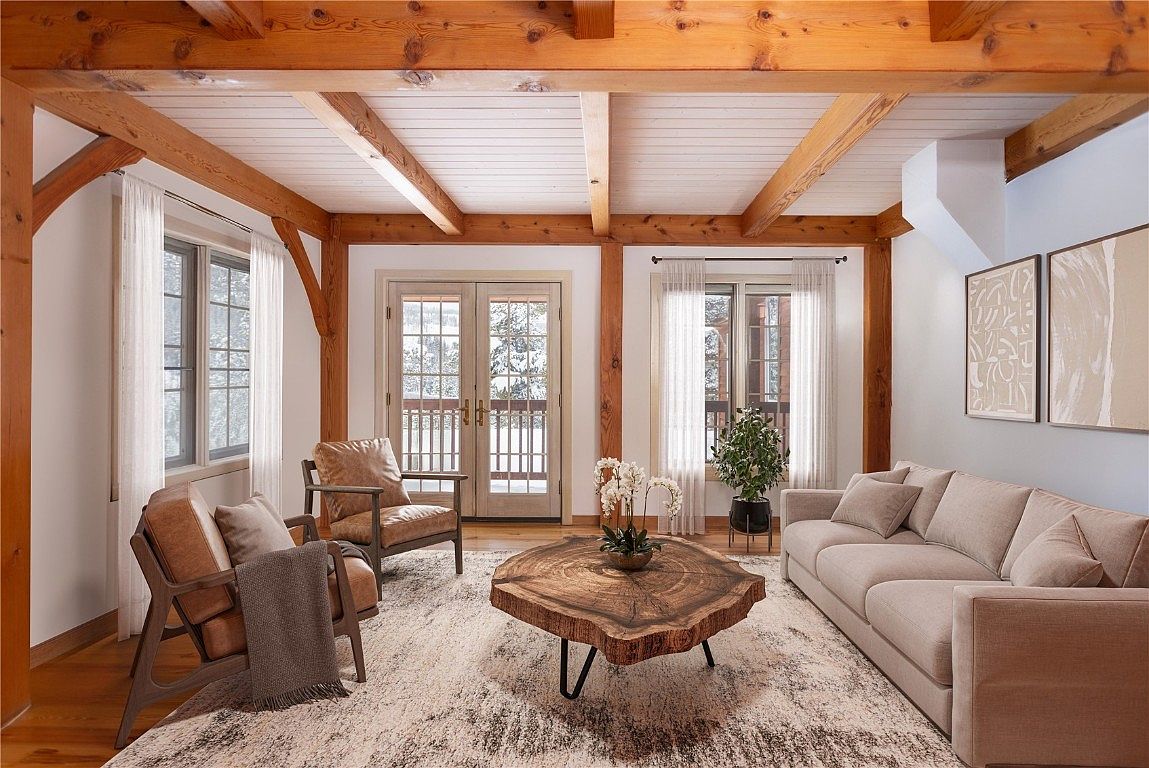 The living room features exposed wooden beams and a light-colored rug, creating a rustic yet inviting atmosphere. The room is furnished with a beige sofa, two leather chairs, and a unique wooden coffee table. Natural light streams in through the windows, enhancing the warm and comfortable ambiance of the space.
