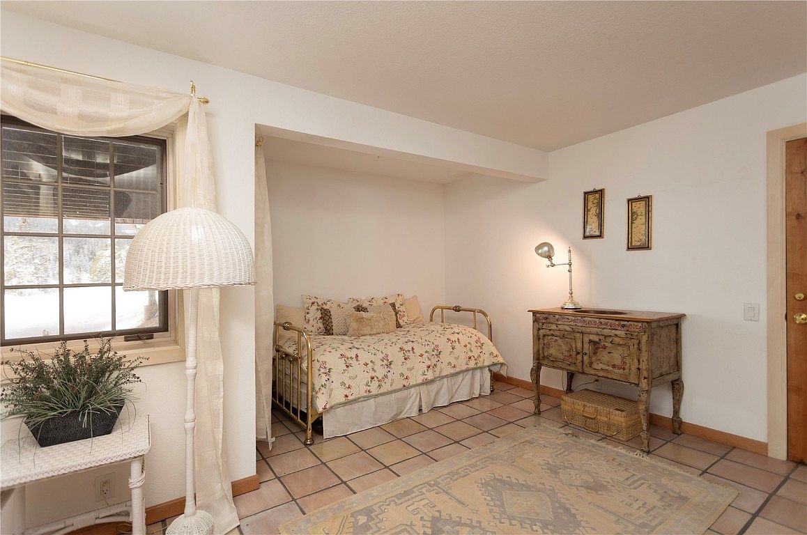 This is an interior shot of a guest bedroom featuring a daybed nestled in an alcove, adorned with floral bedding and decorative pillows. A vintage-style wooden cabinet with a lamp sits beside the bed, complemented by framed artwork on the wall. The room is illuminated by natural light filtering through a window with sheer curtains, creating a cozy and inviting atmosphere.