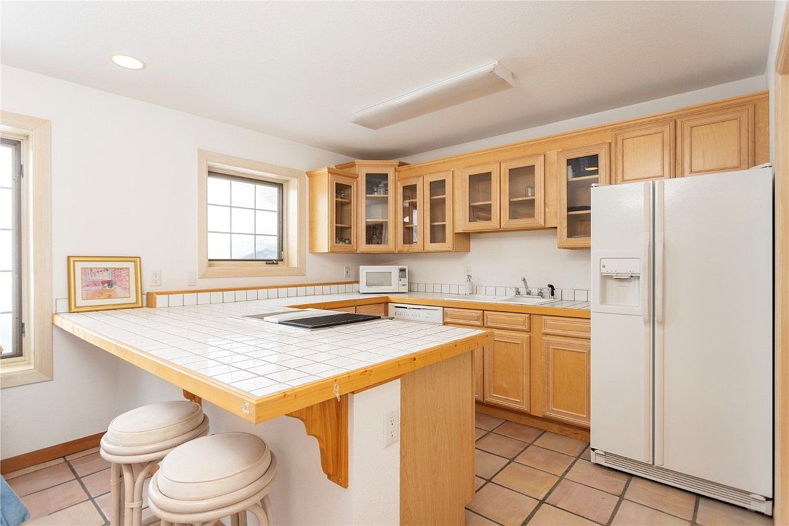 This is a well-lit kitchen featuring light wood cabinetry, a white refrigerator, and a tiled countertop island with seating. The kitchen has a warm, inviting feel with terracotta tile flooring and natural light streaming in from the windows. The perspective is from a medium distance, showcasing the layout and key features of the kitchen.