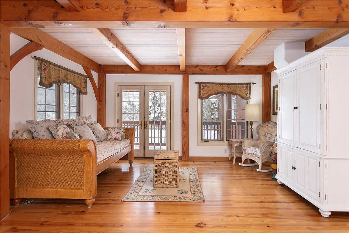 This inviting living room features exposed wooden beams and a light-filled ambiance. A wicker daybed with floral cushions sits beside a window with a floral valance, complemented by a matching wicker chair and a white cabinet. The hardwood floors and area rug add warmth and character to the space.