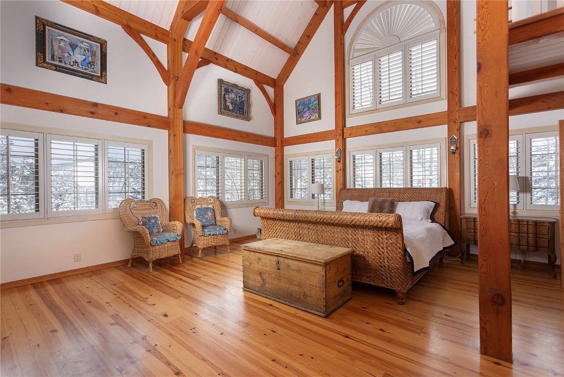 This is a spacious primary bedroom featuring a unique wooden beam structure and a high vaulted ceiling. The room is well-lit with multiple windows and showcases a woven bed frame and a wooden chest. The hardwood floors add warmth to the space, creating a cozy and inviting atmosphere.