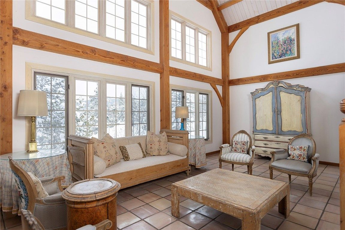 This living room features a blend of rustic and elegant styles. The room is characterized by exposed wooden beams, large windows offering a view of a snowy landscape, and a mix of antique-style furniture including a daybed, armchairs, and a painted cabinet. The tile flooring and area rug add texture and warmth to the space.