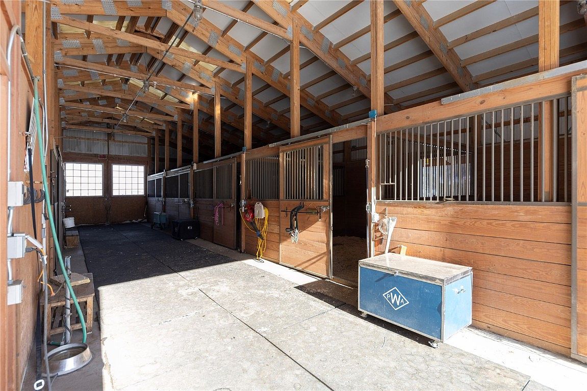 This image showcases the interior of a well-maintained barn, featuring wooden horse stalls with metal bars. The structure has an open, exposed beam ceiling, providing ample height and ventilation. A concrete aisle runs through the center, and a blue storage chest adds a touch of color.
