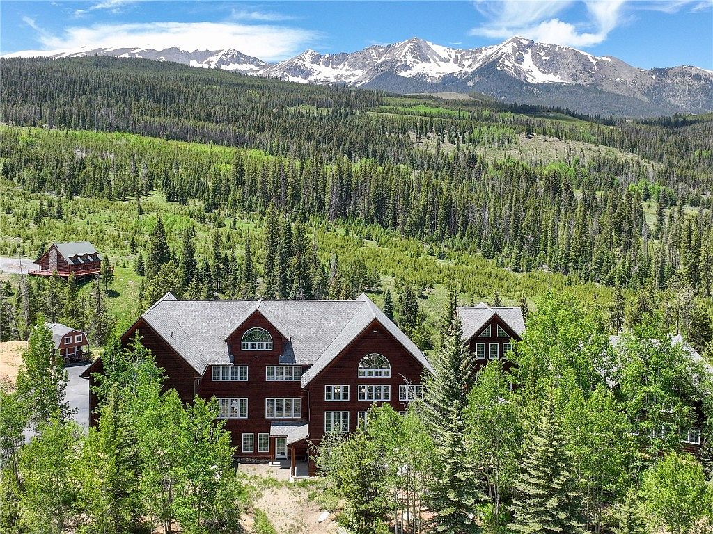 This image showcases the front view of a large, multi-story mountain home with a dark red exterior and gray roof. The house is surrounded by lush green trees and features multiple windows and architectural details. Snow-capped mountains are visible in the background, creating a scenic and desirable setting.