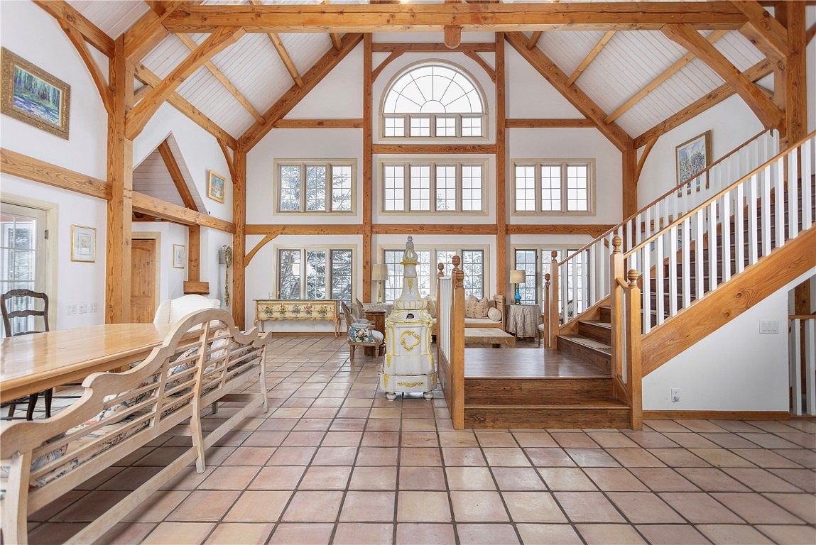 This is a spacious living room featuring exposed wooden beams and a high vaulted ceiling, creating a rustic yet elegant atmosphere. Large windows provide ample natural light, highlighting the tile flooring and the staircase leading to the upper level. The room is furnished with a mix of antique and comfortable seating, suggesting a cozy and inviting space for relaxation and entertaining.