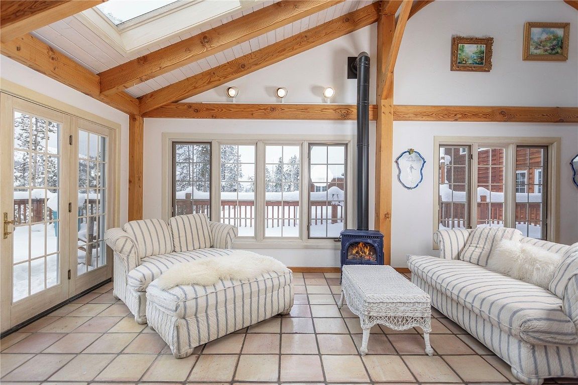 This sunroom features a cozy seating area with striped sofas and a white wicker coffee table, centered around a blue wood-burning stove with a black chimney. Large windows offer views of a snowy landscape, while exposed wooden beams and a skylight add architectural interest. The tiled floor and neutral color palette create a warm and inviting atmosphere.