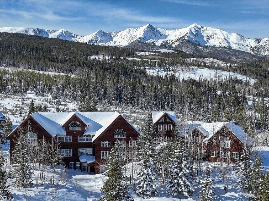 This image showcases the front exterior of a multi-story, chalet-style building with a dark red facade and snow-covered roofs. The property is surrounded by snow-covered ground and evergreen trees, with a backdrop of majestic, snow-capped mountains under a clear blue sky. The scene evokes a sense of luxury and tranquility, ideal for a mountain resort or vacation home.