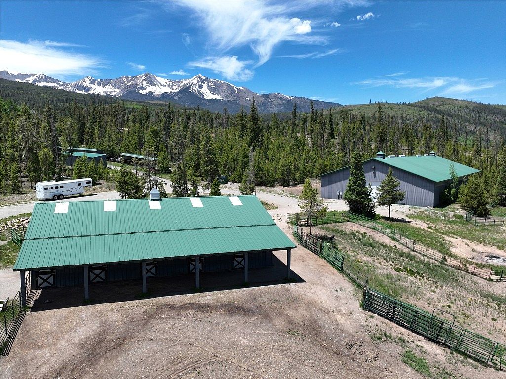 This aerial view showcases a property with multiple buildings, including a large barn-like structure with a green roof and open stalls, a sizable warehouse, and a recreational vehicle. The property is surrounded by a mix of open land and dense forest, with snow-capped mountains visible in the background, creating a picturesque and secluded setting. The perspective is high-angle, providing a comprehensive view of the property's layout and its natural surroundings.