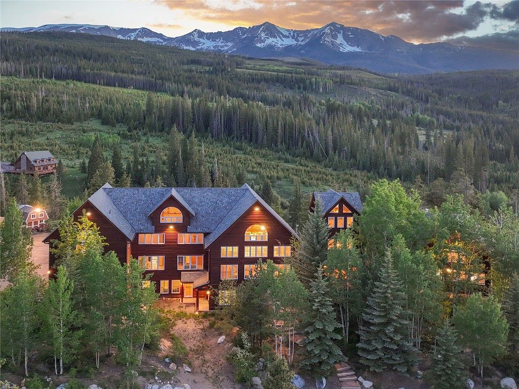This aerial view showcases a grand mountain home nestled among lush evergreen trees, with snow-capped mountains in the background. The home features a dark wood exterior, multiple windows, and a sprawling roofline, exuding a sense of luxury and seclusion. The surrounding landscape emphasizes the property's private and scenic location.