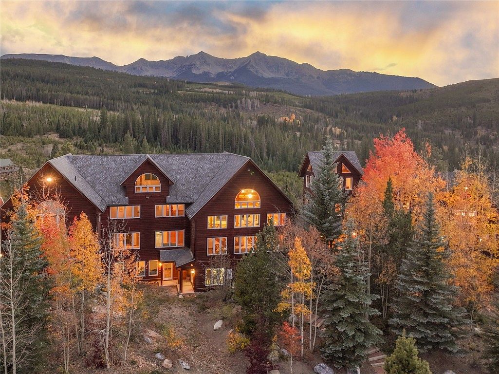 This image showcases the front exterior of a large, multi-story mountain home with a rustic, lodge-like design. The home features a dark wood facade, a gray shingle roof, and numerous windows that suggest ample natural light within. The surrounding landscape includes colorful autumn trees and a backdrop of forested mountains, creating a serene and picturesque setting.