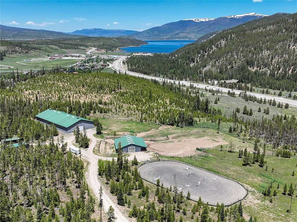 This aerial shot showcases a sprawling property featuring a large barn with a green roof, a smaller building, and an outdoor riding arena. The property is surrounded by lush greenery and trees, with a lake and mountains visible in the background. A road runs alongside the property, adding to the overall sense of space and accessibility.