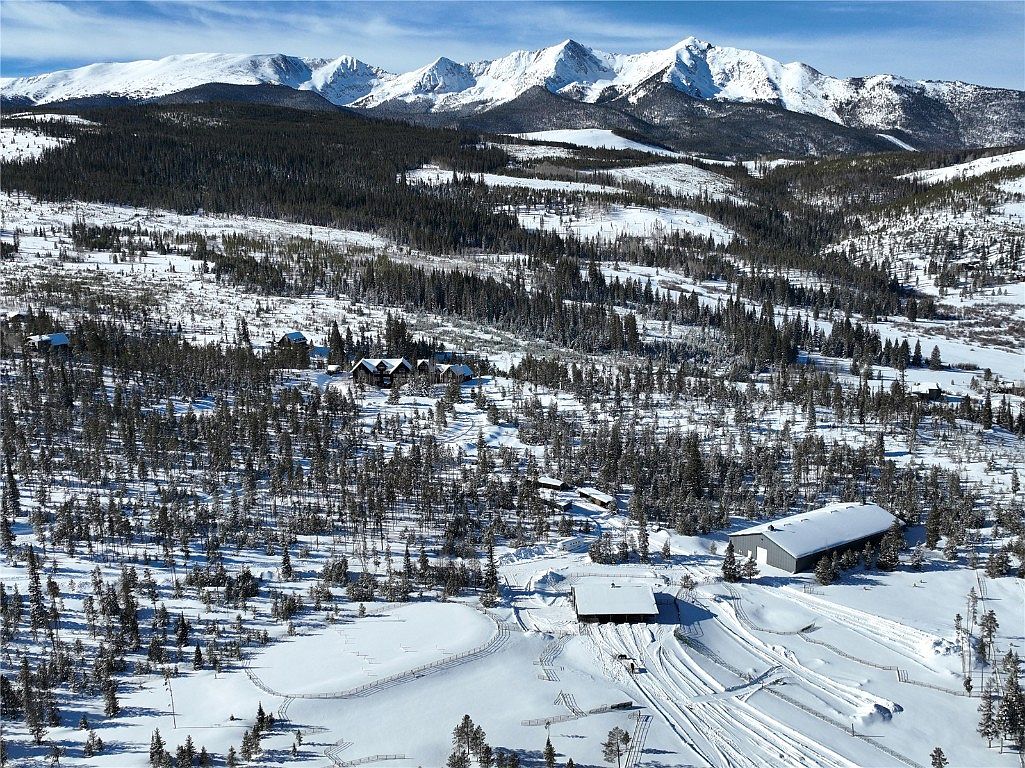 This aerial view showcases a sprawling estate nestled in a snow-covered mountain landscape. The property features multiple buildings, including a large barn-like structure and several residential homes, all connected by snow-covered pathways. The backdrop of snow-capped mountains and dense evergreen forests creates a serene and secluded atmosphere, highlighting the property's privacy and natural beauty.