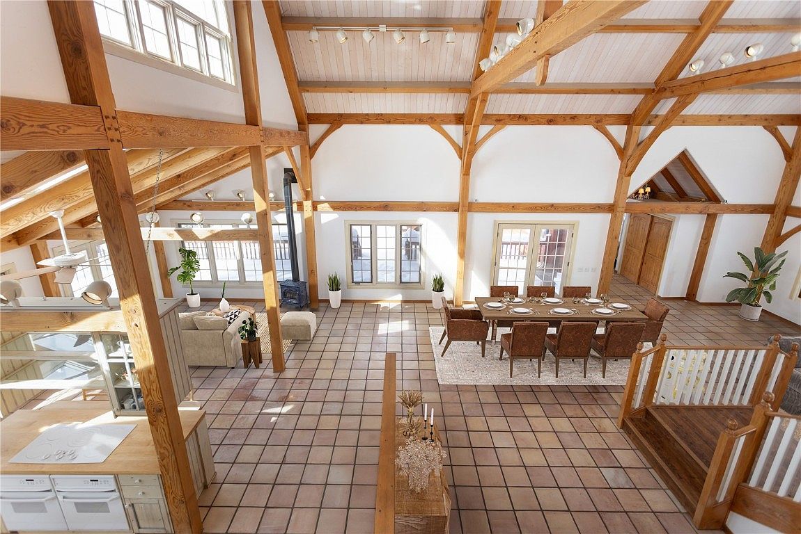This is a spacious living area with exposed wooden beams and a high ceiling, creating a rustic yet modern atmosphere. The room features a dining table set for eight, a cozy seating area with a sofa and ottoman, and a fireplace. Large windows provide ample natural light, highlighting the open floor plan and tiled flooring.