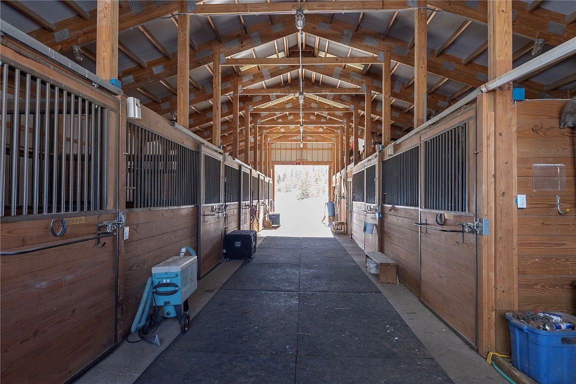 This image showcases the interior of a barn, featuring wooden stalls with metal bars along both sides of a central corridor. The structure has a high, exposed wooden beam ceiling with lighting fixtures. The floor is covered with a dark mat, and the open end of the barn reveals an outdoor scene with trees and natural light.