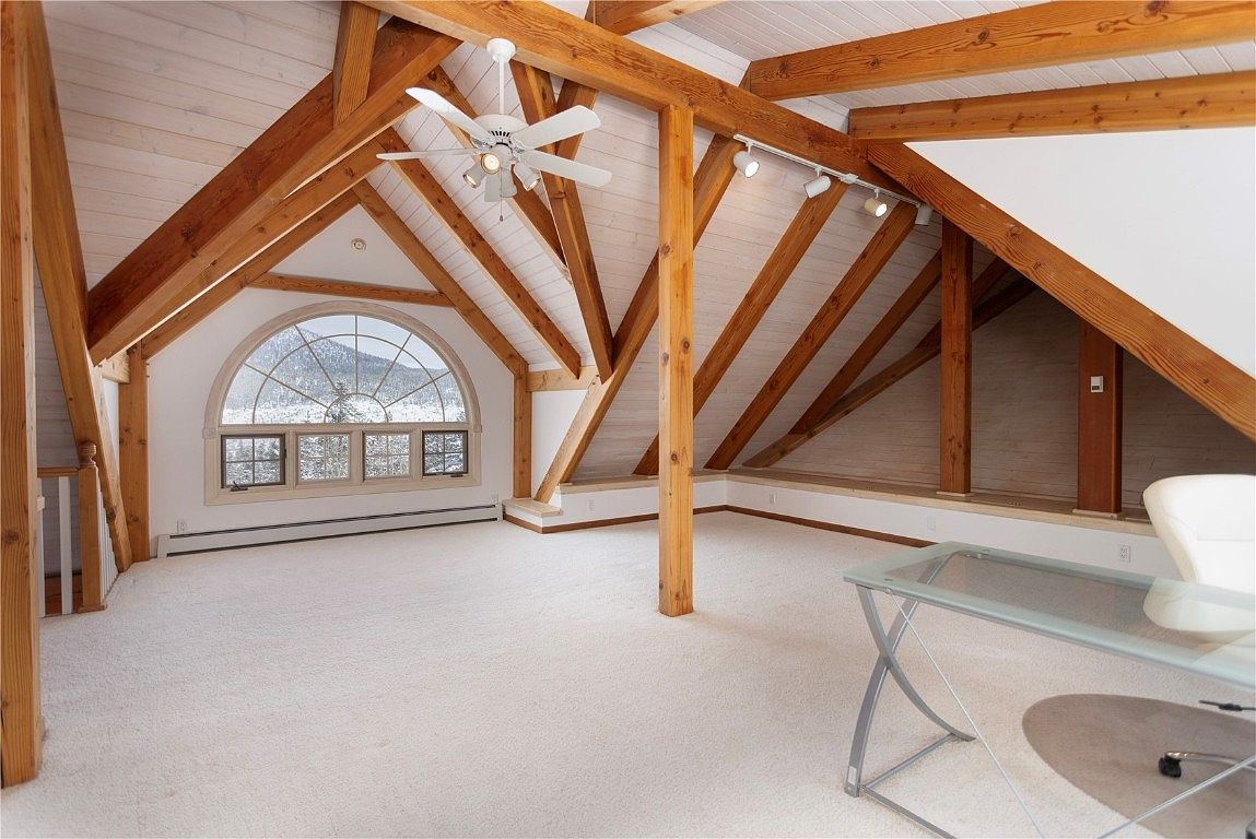 This is an interior shot of a spacious attic, featuring exposed wooden beams and a vaulted ceiling. A large arched window provides natural light and a view of the outdoors. The room is carpeted in a light color, creating a bright and airy atmosphere, and a modern desk is visible in the foreground.