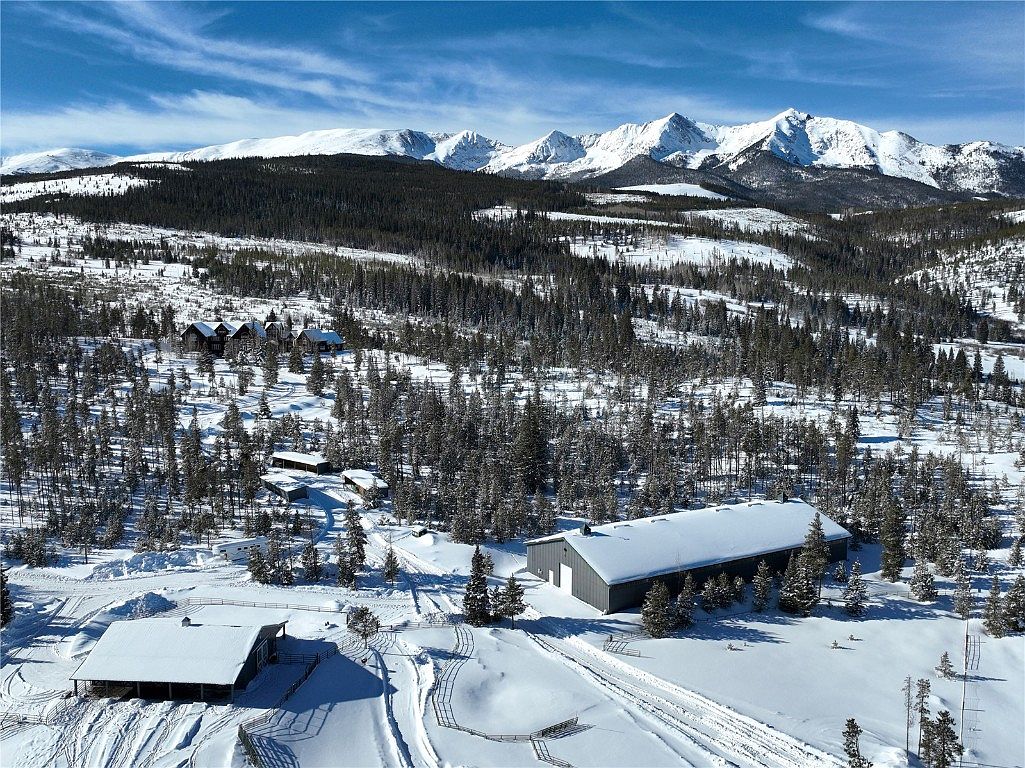 This aerial view showcases a sprawling property nestled in a snowy, mountainous landscape. Several buildings, including a large barn-like structure and smaller cabins, are visible amidst the snow-covered trees. The majestic snow-capped mountains in the background add a sense of grandeur and seclusion, highlighting the property's unique setting.