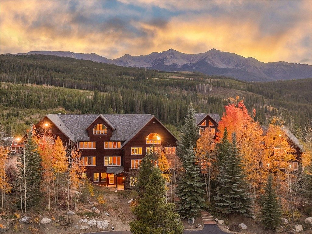This image showcases the front exterior of a large, multi-story mountain lodge-style home. The building features a dark wood facade, multiple windows with warm interior lighting, and a gray shingle roof. The surrounding landscape includes vibrant autumn foliage and evergreen trees, with a backdrop of distant mountains under a colorful sky, creating a picturesque and inviting scene.
