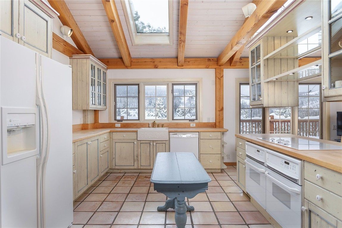 This is a kitchen with rustic charm, featuring light-colored wooden cabinets, butcher block countertops, and a farmhouse sink. The room is well-lit by natural light from multiple windows, offering views of a snowy landscape. A central island provides additional workspace, and the tiled floor adds to the cozy, country feel.