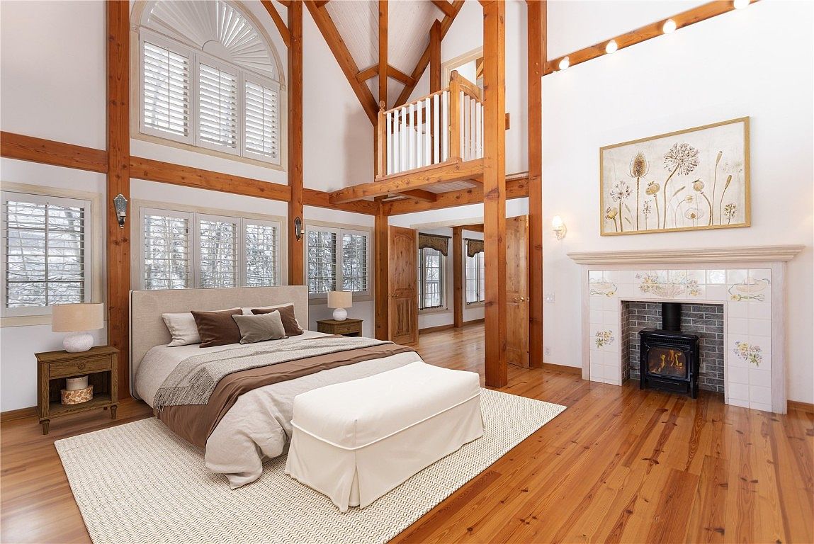 This is a spacious primary bedroom featuring exposed wooden beams and a high ceiling with a loft area. The room includes a large bed with neutral-toned bedding, a fireplace with decorative tiles, and large windows with shutters. The hardwood floors and area rug add warmth and texture to the space, creating a cozy and inviting atmosphere.
