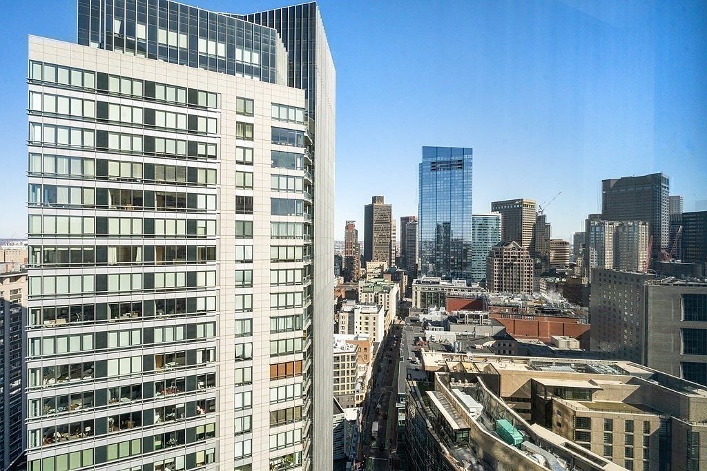This high-angle aerial view captures a dense urban cityscape, dominated by a prominent, modern high-rise building with a grid-like facade of glass and light-colored panels. The perspective looks down onto a bustling street canyon, revealing a mix of contemporary skyscrapers and older brick structures. The clear blue sky provides a bright, expansive backdrop, emphasizing the scale and density of the metropolitan environment.