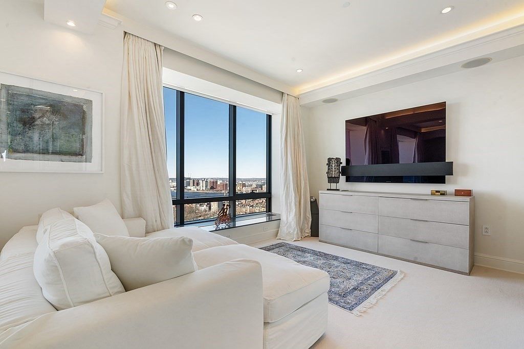 This bright and airy living room features a plush white sofa positioned to take advantage of the large windows that offer a panoramic city view. A sleek, light-toned dresser serves as a media console beneath a wall-mounted television, while a patterned area rug adds texture to the neutral carpeted floor. The space is finished with elegant floor-length curtains and recessed lighting, creating a sophisticated and serene atmosphere.