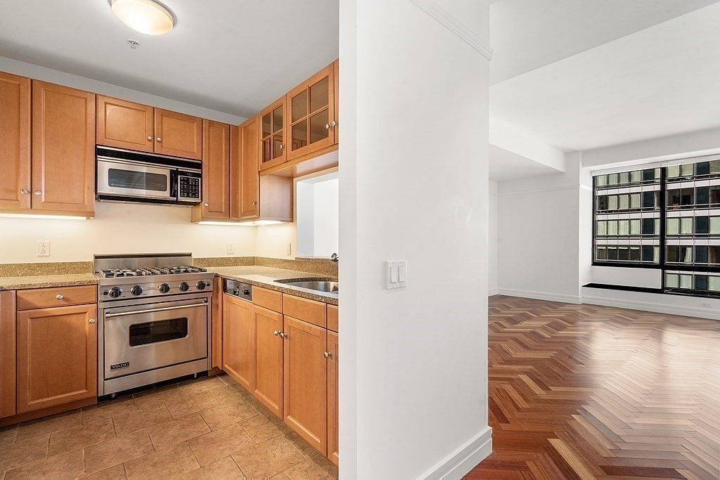 This kitchen features light wood cabinetry, a stainless steel Viking range, and a built-in microwave, all set against a neutral backsplash and stone countertops. The space is tiled with light-colored flooring and transitions into an adjacent living area with dark, herringbone-patterned wood floors. The perspective is from the kitchen entrance, looking toward the living room, which is illuminated by large windows.