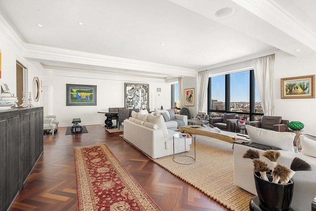 This expansive, open-concept living area features rich, dark herringbone-patterned wood flooring and a neutral color palette that emphasizes a sophisticated, airy atmosphere. The space is anchored by a large, textured area rug and plush white sofas, complemented by a natural wood coffee table and a long, dark console cabinet. Large windows provide an abundance of natural light and expansive city views, while curated artwork and decorative accents add a refined, gallery-like aesthetic to the room.