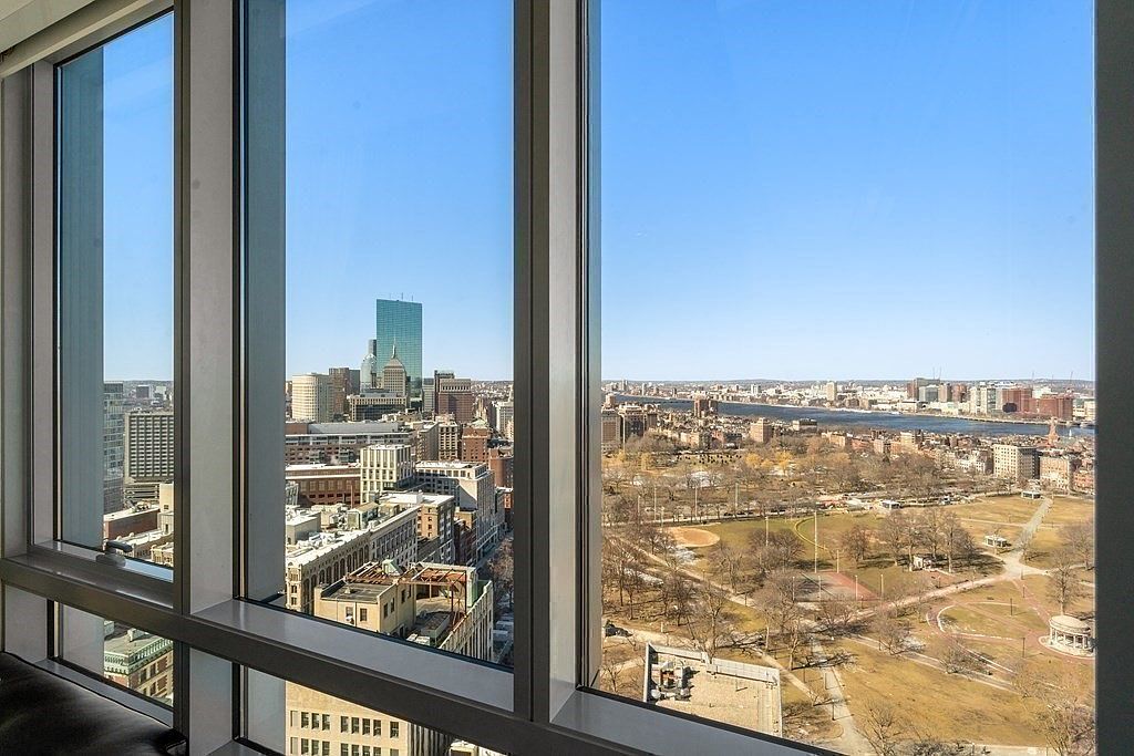 This interior view features expansive floor-to-ceiling windows that frame a panoramic cityscape, including a prominent skyscraper and a large public park. The perspective is from a high-rise vantage point, looking out over the urban landscape and a river in the distance. The clean, modern framing of the windows emphasizes the dramatic height and the impressive view, creating a bright and airy atmosphere.