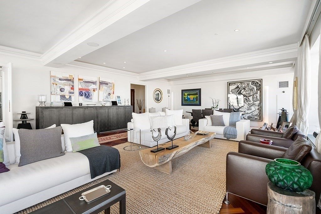 This expansive, light-filled living room features a sophisticated open-concept layout with white sofas, leather armchairs, and a unique live-edge wood coffee table. The space is decorated with contemporary art pieces and a large, textured area rug, creating a refined yet comfortable atmosphere. The perspective captures the depth of the room, showcasing the transition from the seating area toward a dining space in the background.