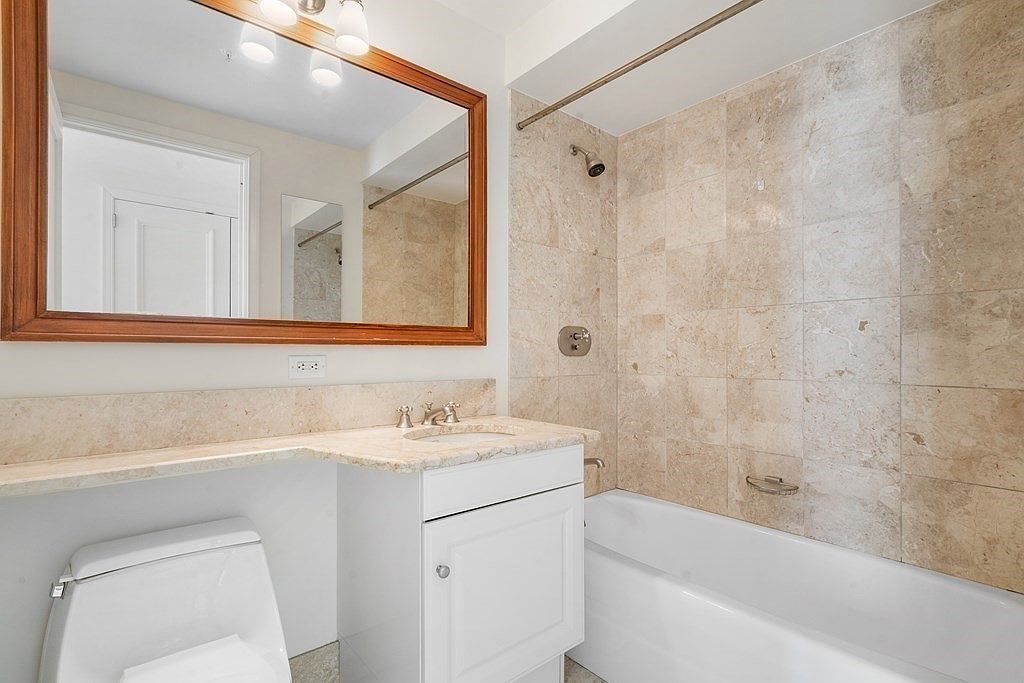 This compact bathroom features a white vanity with a stone countertop and an integrated sink, paired with a large, wood-framed mirror. The shower-tub combination is enclosed by light-toned, square-patterned wall tiles that extend to the ceiling. The space is bright and functional, utilizing a neutral color palette to maximize the sense of openness in a smaller footprint.