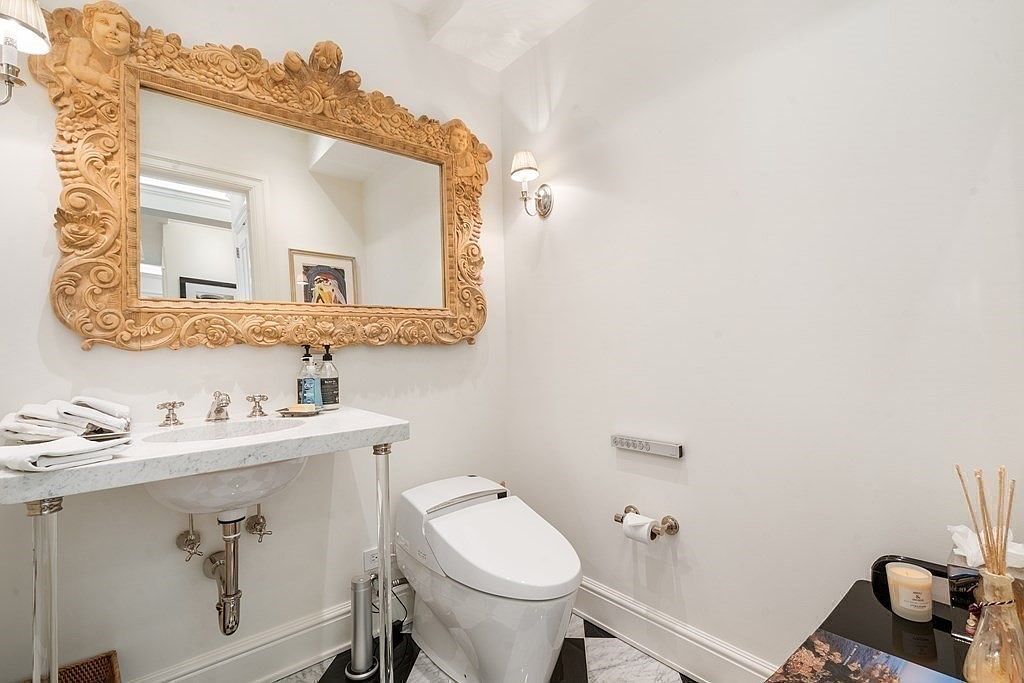 This elegant powder room features a striking ornate gold-framed mirror mounted above a wall-mounted stone vanity with clear acrylic legs. The space is finished with a black-and-white checkered floor and a modern white toilet, creating a sophisticated and high-contrast aesthetic. A wall-mounted light fixture provides warm illumination, while a small side table holds decorative accents, adding a refined touch to the compact room.