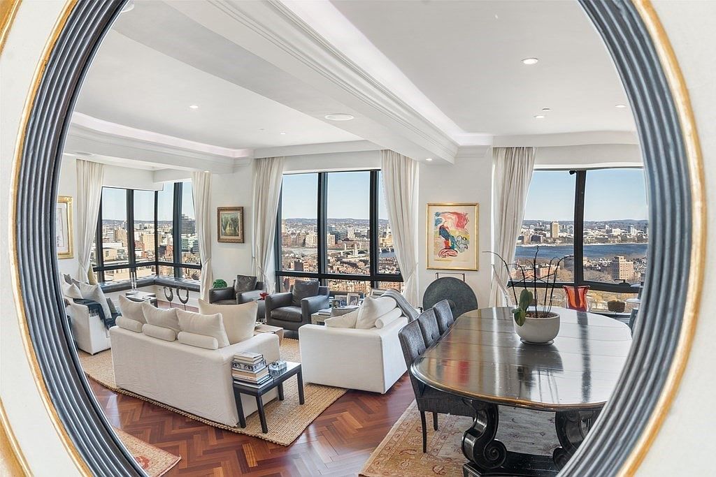 This image is captured through a large, circular mirror, providing a unique, wide-angle perspective of an elegant open-concept living and dining area. The space features rich, dark herringbone wood flooring, plush white sofas, and expansive windows that offer a panoramic view of the city skyline and river. The interior design is sophisticated and airy, with neutral tones, framed artwork, and a formal dining table positioned to take advantage of the natural light.