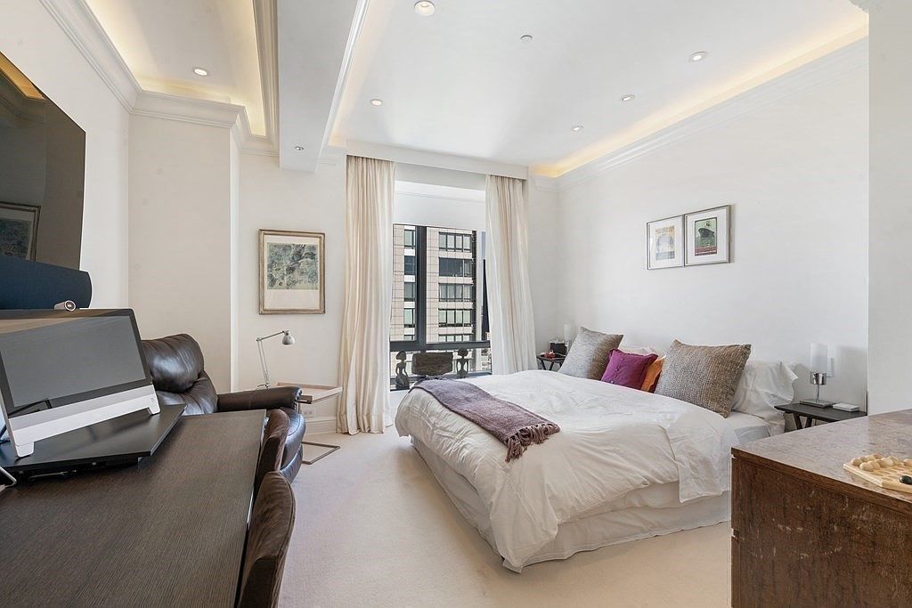 This primary bedroom features a neutral color palette with plush carpeting and a large bed centered against a wall adorned with two framed prints. To the left, a dark wood desk and leather armchair create a functional workspace, while tall windows draped in floor-length curtains allow natural light to fill the room. The space is finished with recessed lighting and a tray ceiling, offering a sophisticated and tranquil atmosphere.