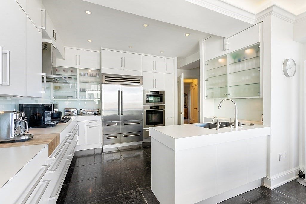 This modern, high-end kitchen features sleek white cabinetry, stainless steel appliances, and a central island with a built-in sink. The space is characterized by its clean lines, dark stone flooring, and glass-fronted upper cabinets that add a sense of openness. A wide-angle perspective captures the functional layout, highlighting the professional-grade refrigerator and wall-mounted ovens.