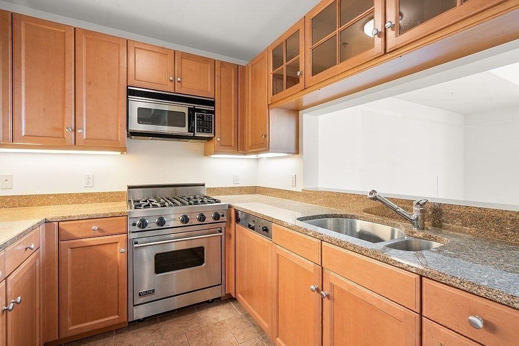 This kitchen features light wood cabinetry, speckled stone countertops, and stainless steel appliances, including a professional-style gas range. A pass-through window above the sink provides an open view into the adjacent room, enhancing the sense of space. The layout is efficient and functional, with under-cabinet lighting illuminating the workspace.