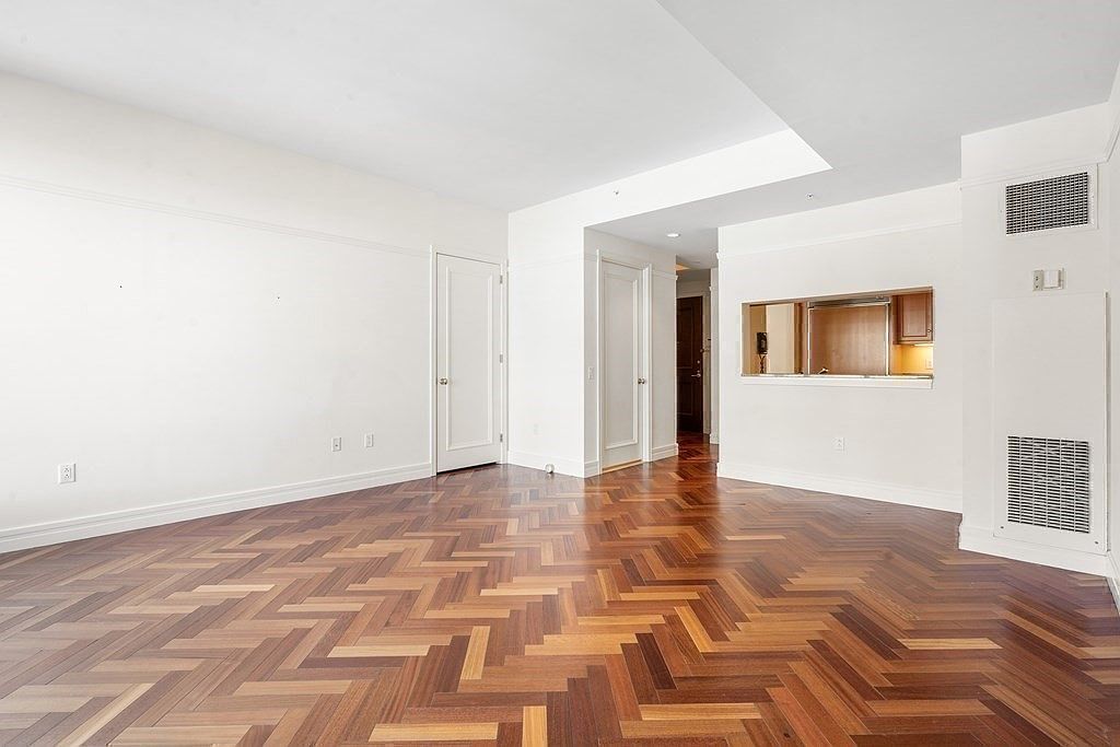 This bright, open-concept living space features rich, dark wood flooring laid in a classic herringbone pattern. The room is characterized by crisp white walls, clean lines, and a pass-through window that offers a glimpse into the adjacent kitchen area. The perspective is captured from a low angle, emphasizing the expansive floor space and the transition into the hallway beyond.