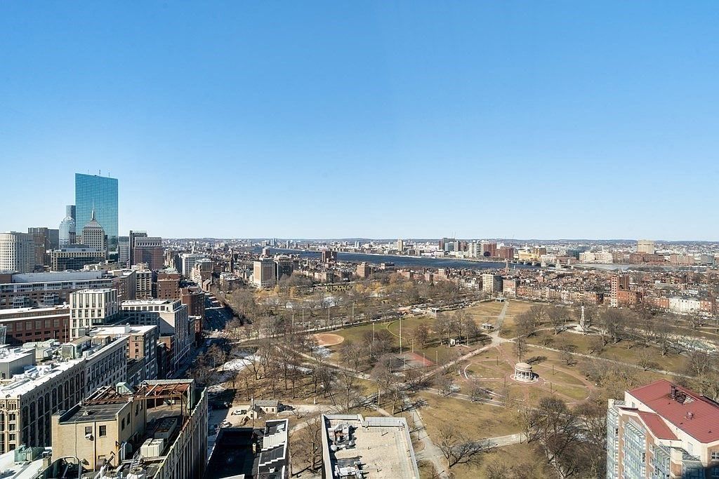 This high-angle aerial view captures a sprawling urban landscape, prominently featuring a large public park with winding paths and bare trees in the foreground. Beyond the park, the dense city skyline rises, highlighted by the iconic, tall, blue-glass skyscraper, with the Charles River stretching horizontally across the mid-ground. The perspective offers a comprehensive look at the city's layout, emphasizing the proximity of green space to the urban core under a clear, bright blue sky.