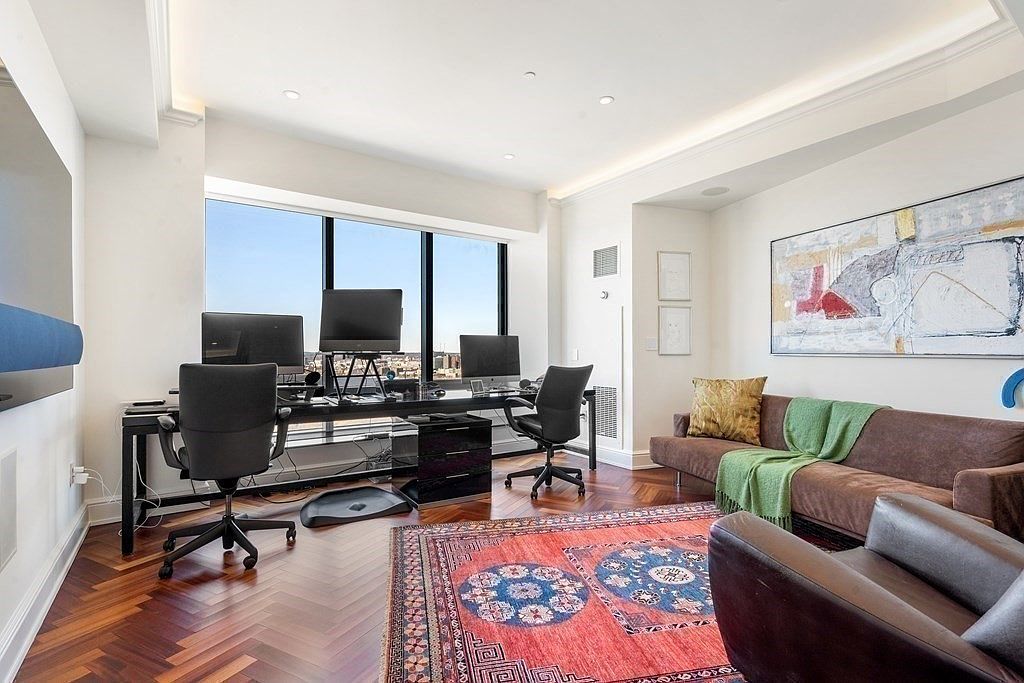 This sophisticated home office features rich, dark wood herringbone flooring and a large desk setup positioned in front of expansive windows that offer a panoramic city view. The room is furnished with a brown sofa adorned with a green throw blanket, a leather armchair, and a vibrant, patterned area rug that adds warmth to the space. A large piece of abstract art hangs on the wall, contributing to the professional yet comfortable atmosphere of this well-lit workspace.
