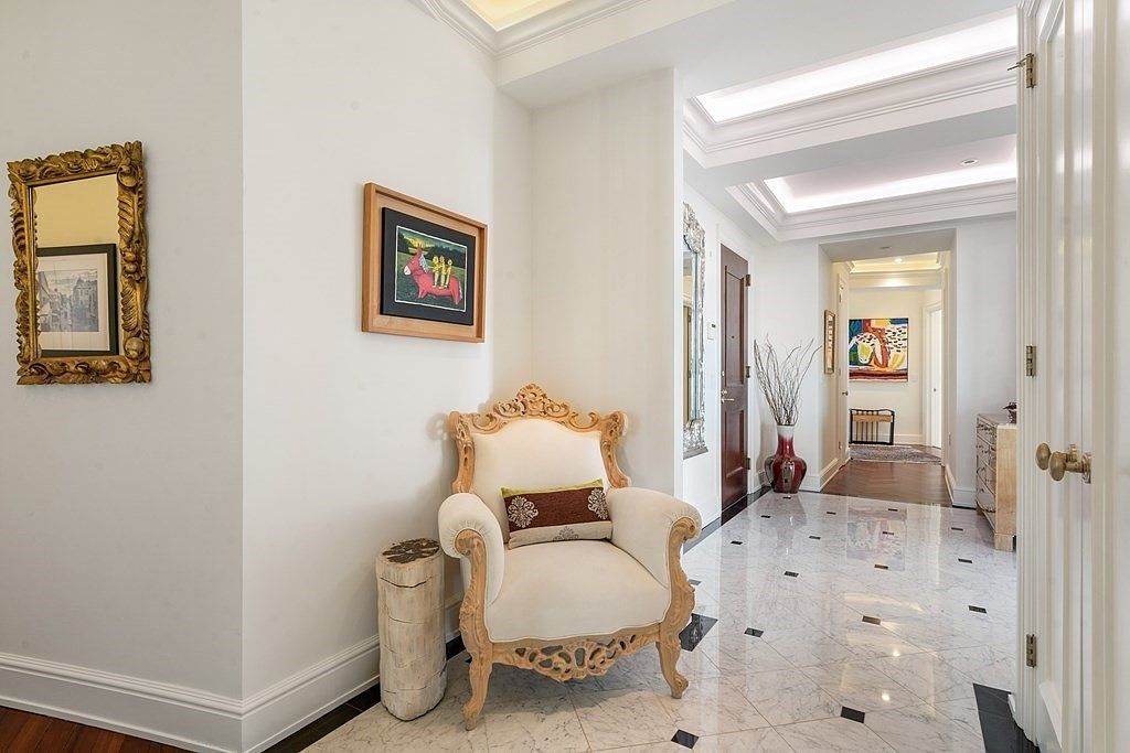 This elegant foyer features polished stone flooring with dark accent inlays, leading into a bright, gallery-style hallway. The space is furnished with a classic, ornate armchair and a rustic log-style side table, complemented by framed artwork and a decorative gold-framed mirror. The perspective looks from the entryway into a well-lit corridor, highlighting the sophisticated architectural details and recessed ceiling lighting.