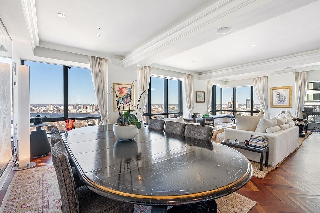 This elegant open-concept living and dining space features a large, dark-toned oval dining table as the centerpiece, surrounded by upholstered chairs. The room is bathed in natural light from expansive windows that offer panoramic city and waterfront views, complemented by a neutral color palette and rich herringbone-patterned wood flooring. A plush white sofa and seating area are visible in the background, creating a sophisticated and airy atmosphere suitable for high-end urban living.