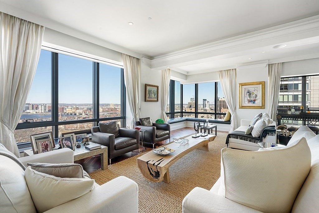 This sophisticated living room features a bright, open layout with plush white sofas and dark leather armchairs arranged around a unique, live-edge wood coffee table. The space is defined by expansive, floor-to-ceiling windows that offer a panoramic urban skyline view, complemented by light-colored curtains and a textured area rug. The room is decorated with framed artwork and personal accents, creating a refined and welcoming atmosphere.