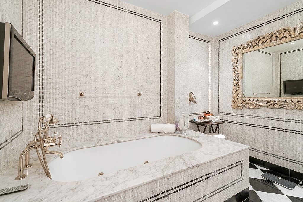 This luxurious primary bathroom features an elegant soaking tub encased in light-colored stone with intricate detailing. The walls are adorned with small, neutral-toned mosaic tiles framed by dark decorative borders, complemented by a large, ornate gold-framed mirror. The space is finished with a striking black and white checkered floor, creating a sophisticated and opulent atmosphere.
