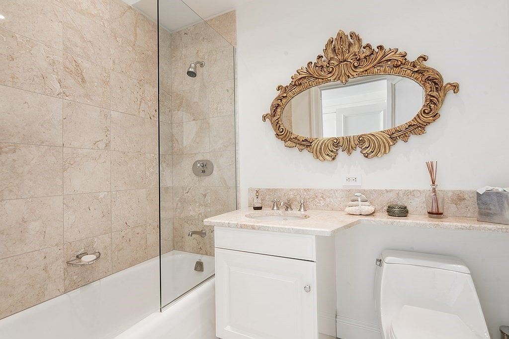 This bright bathroom features a combination tub and shower enclosed by a glass panel, with light-toned stone tile walls. A vanity with a stone countertop sits beside a white toilet, complemented by an ornate, gold-toned decorative mirror mounted on the wall. The space is clean and well-lit, offering a classic and elegant aesthetic.