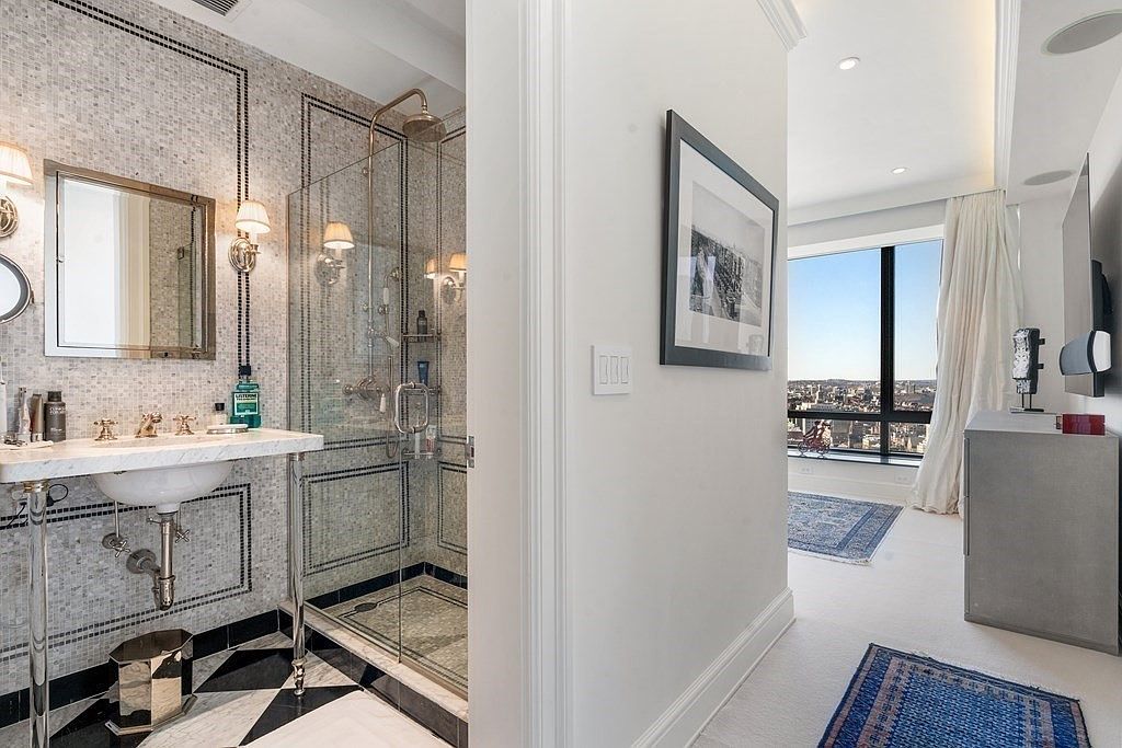 This image captures a sophisticated transition between a luxurious bathroom and an adjacent bedroom. The bathroom features a glass-enclosed shower with intricate mosaic wall tiles, a pedestal sink with a polished metal frame, and a classic black-and-white patterned floor. Beyond the doorway, the bedroom is bathed in natural light from large windows that offer an expansive urban view, complemented by a soft rug and a minimalist aesthetic.