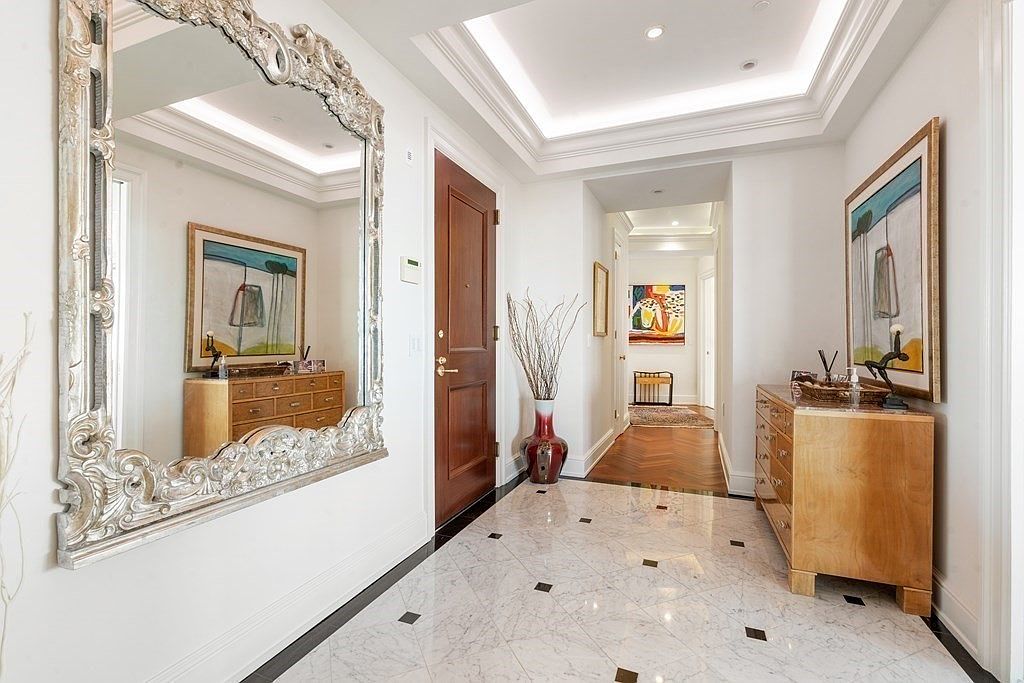 This elegant foyer features a polished light-toned stone floor with dark accent inlays, creating a sophisticated entry point. A large, ornate silver-framed mirror reflects the space, while a wooden console table and a tall vase with decorative branches add texture and warmth. The perspective looks down a bright, recessed-lit hallway that transitions into a rich wood-floored corridor, showcasing a clean and upscale transitional design.