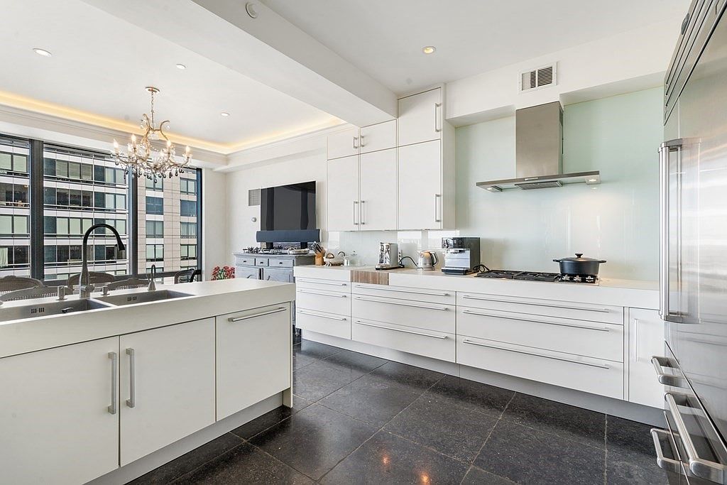 This modern, high-end kitchen features sleek white cabinetry, a large central island with a double sink, and dark stone flooring. The space is equipped with professional-grade stainless steel appliances, including a built-in coffee machine and a range hood, all set against a light-colored backsplash. Large windows provide an expansive view of the surrounding urban architecture, while a crystal chandelier adds a touch of elegance to the bright, open-concept layout.
