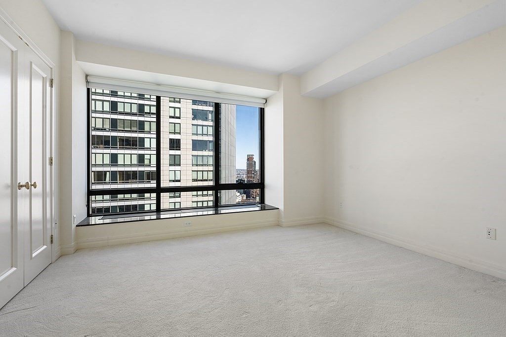 This bright, minimalist bedroom features neutral carpeting and crisp white walls, creating a clean and versatile living space. A large window wall provides an expansive view of the surrounding city skyline, allowing for significant natural light. The room includes a simple, functional layout with a dark stone sill beneath the windows and a set of white doors on the left.